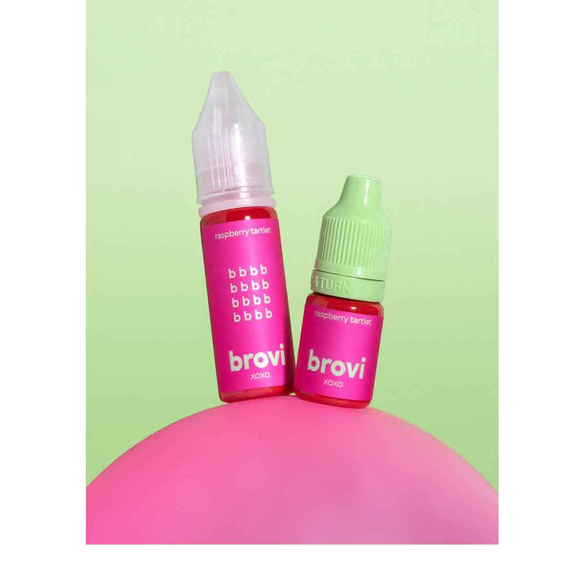 Two Brovi e-liquid bottles labeled 'raspberry basket' in pink, one with a clear cap and one with a mint green cap, resting on a pink surface against a light green background.