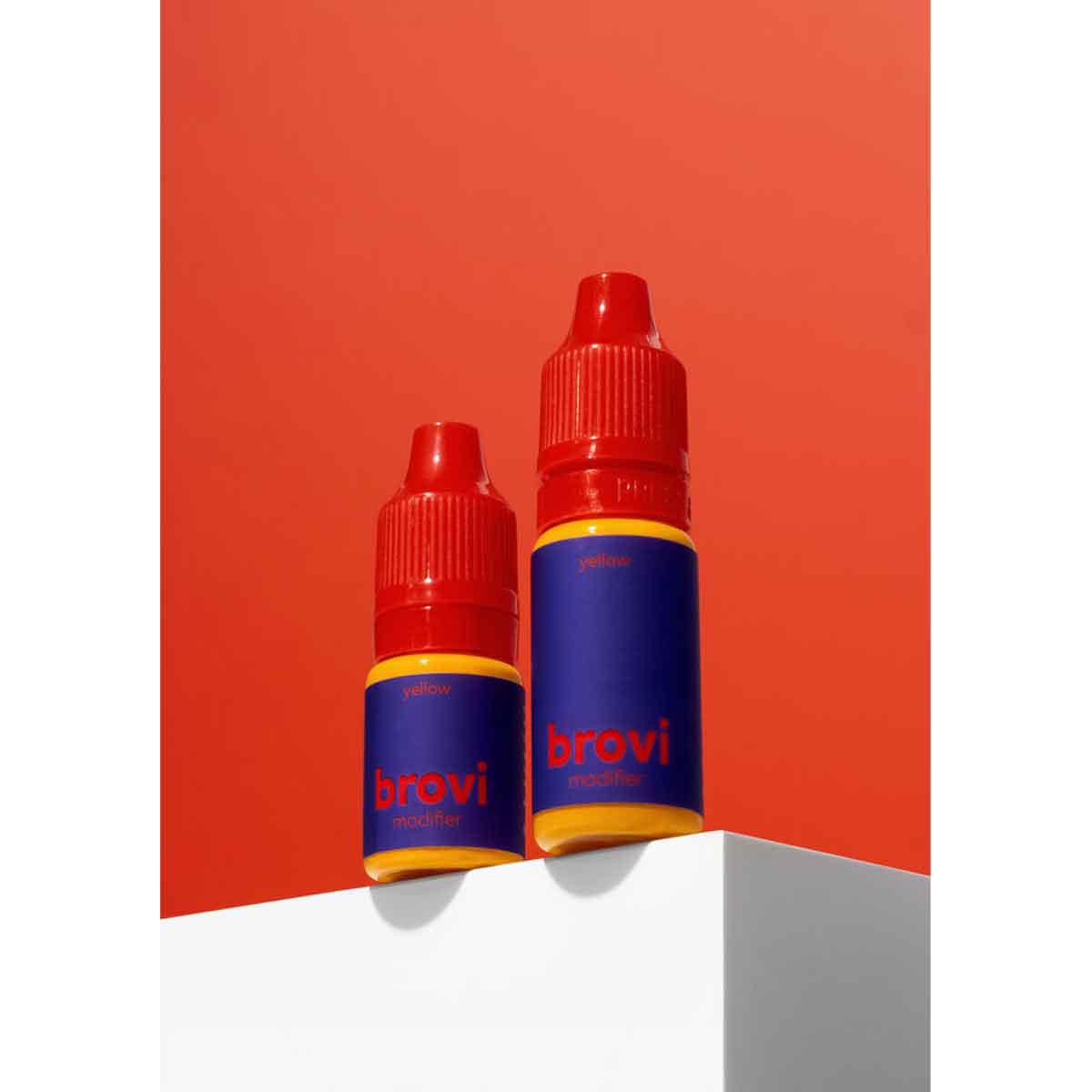 Two Brovi nicotine e-liquid bottles with red caps and blue labels, displayed on a white geometric pedestal against a solid red-orange background.