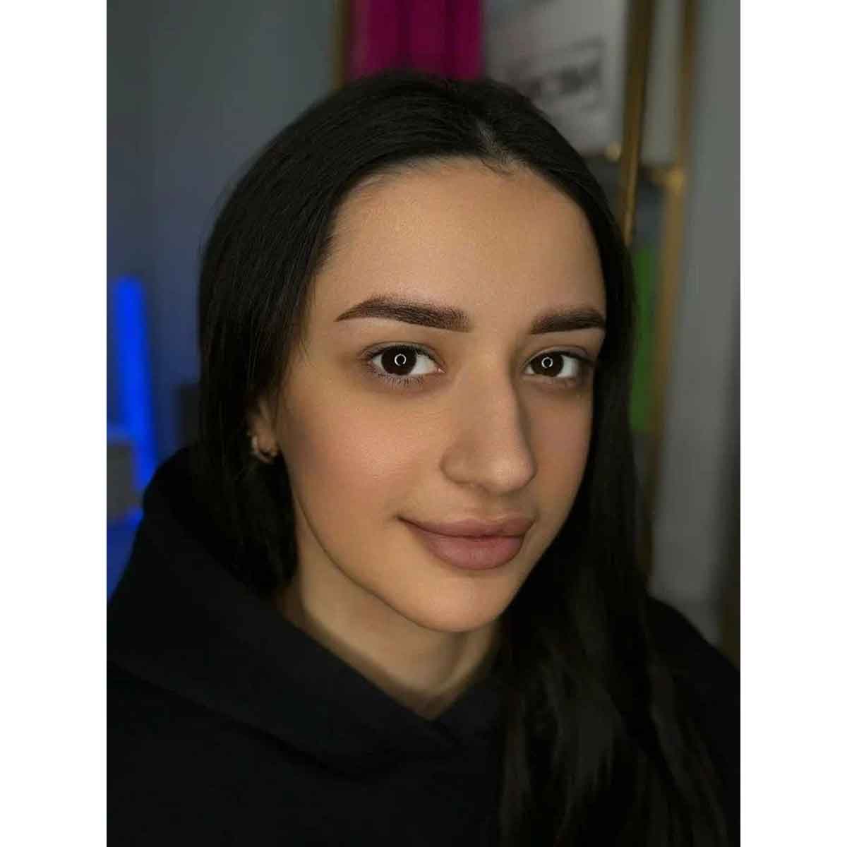 A close-up portrait of a woman with long dark hair wearing a black hoodie, set against an indoor background with soft, out-of-focus colors.