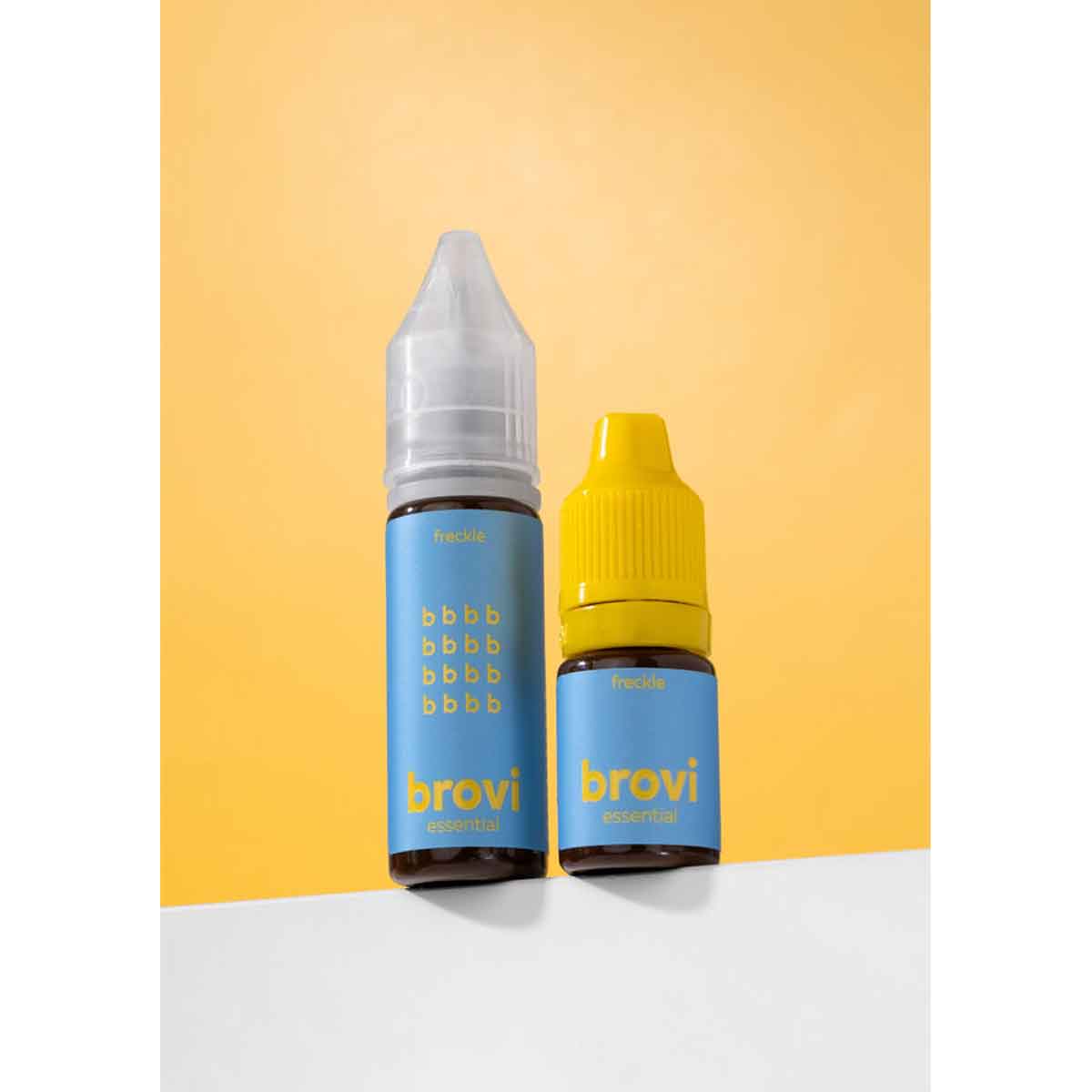 Two Brovi essential oil dropper bottles, one with a clear cap and one with a yellow cap, standing on a white surface against a yellow background.