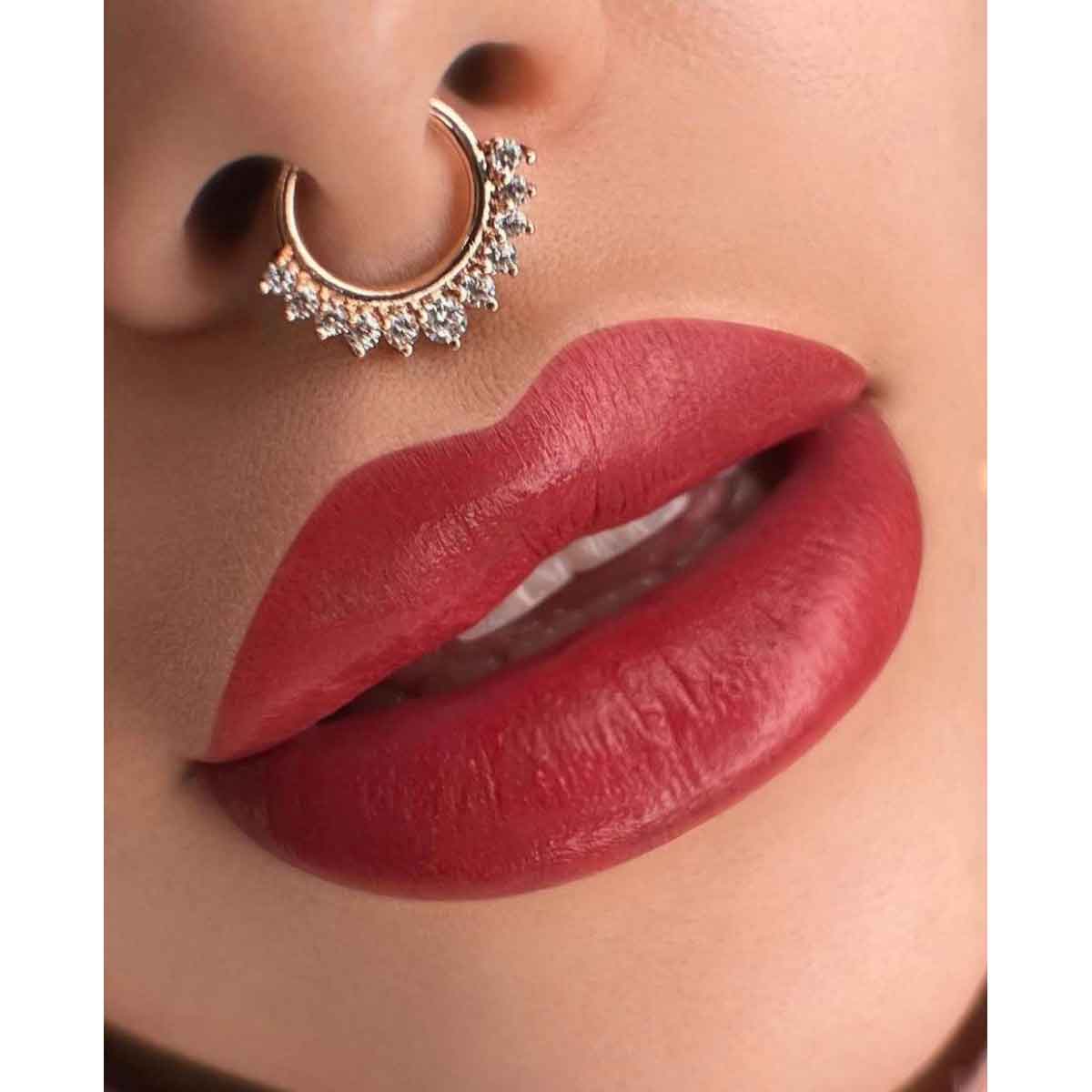 A close-up of a person's face wearing a gold crescent-shaped nose hoop adorned with small clear rhinestones, paired with glossy red lipstick.