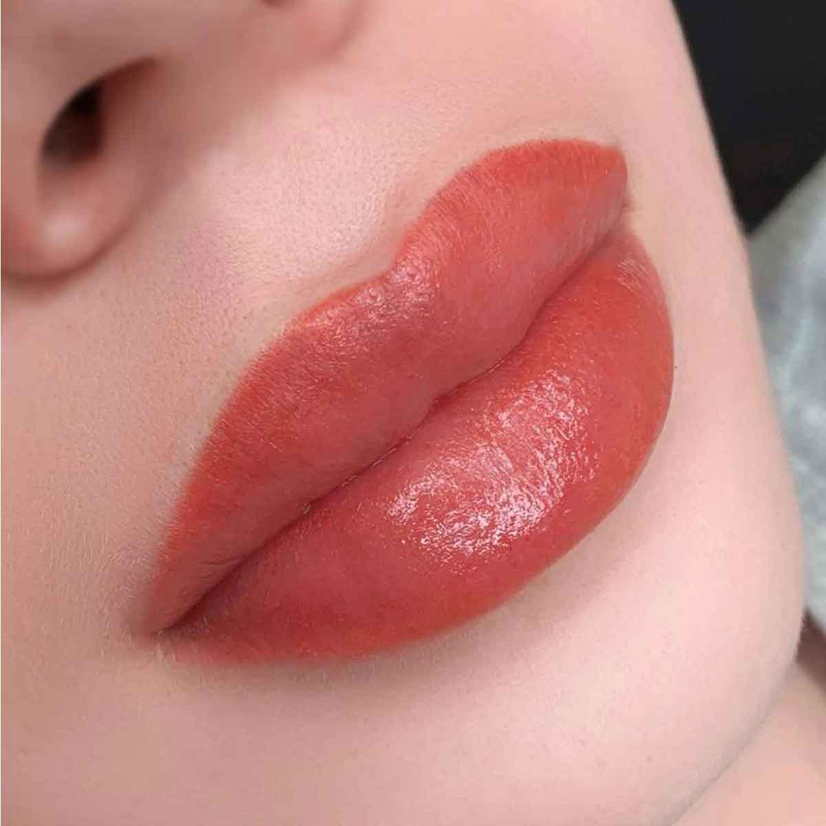 Close-up of a person's lips wearing a glossy, warm coral-red lip color.